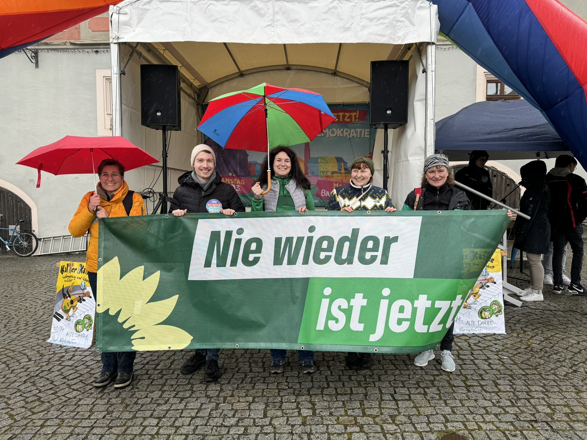 Gruppenbild von Mitgliedern der GRÜNEN in Rhön-Grabfeld, sie halten ein Spruchband in grüner Parteioptik mit der Aufschrift "Nie wieder ist jetzt", im Hintergrund eine Bühne und eine aufblasbarer Regenbogen